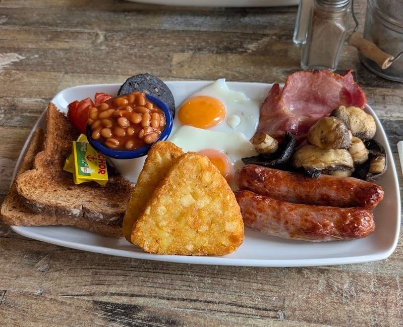 A full English breakfast on a white plate: toast, beans in a pot, two fried eggs, bacon, sausages, mushrooms and a hash brown