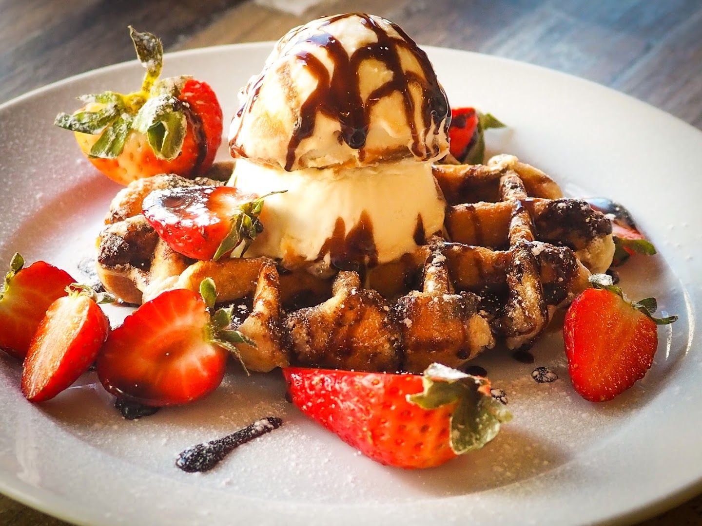 A waffle topped with vanilla ice cream and chocolate sauce, ringed with halved strawberries and dusted with icing sugar